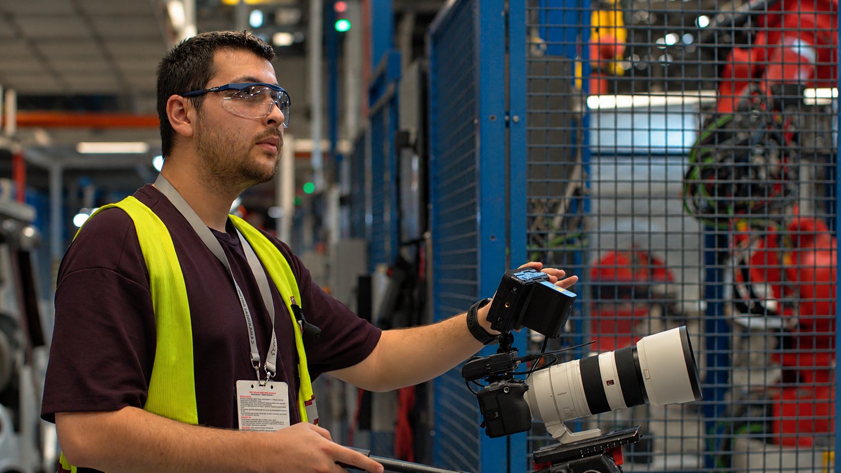 Camera crew member in a safety vest operating a camera on the Tofaş factory floor