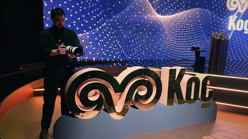 Sefa checking a camera on stage beside a large Koç logo